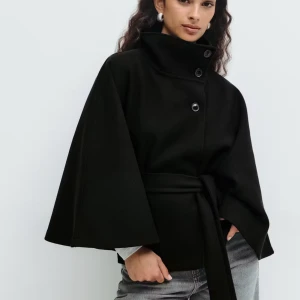 Snygg poncho kappa - Svart kappa med hög krage, stora knappar och bälte i midjan. Jackan har vida ärmar och en stilren, minimalistisk look. Perfekt för dig som gillar en clean och modern stil. Från Gina. Säljer för råka köpa två 😬❤️Skriv innan köp!!❤️