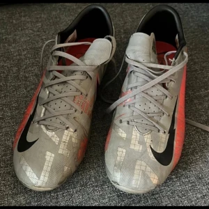 Nike Mercurial grå fotbollsskor - Nike Mercurial fotbollsskor i grått med svarta och röda detaljer. Snyggt mönstrad ovandel med stora reflekterande bokstäver och klassisk svart swoosh. Skorna har snörning och platt sula, insida i rött och svart. Perfekt för dig som vill sticka ut på planen.