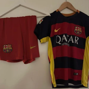 FC Barcelona Messi 10 matchtröja Nike - Barça fotbollströja med Messi 10-tryck, officiell Nike Dri-FIT. Röd och mörkblå med gula detaljer, klubbmärke och Qatar Airways-logga. Korta ärmar, randigt mönster och tillhörande röda shorts med klubbmärke och Nike-logga. Perfekt för fans av FC Barcelona.