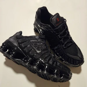 Nike Shox svarta sneakers - Svarta Nike Shox sneakers med ikonisk fjädrande sula och meshdetaljer. Skorna har snörning, diskreta röda detaljer på plösen och bak, samt en sportig vibe. Ovandelen är i syntet och mesh för extra andningsförmåga och komfort.