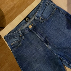 Lee Scarlett High blå jeans W30 L33 - Snygga blå jeans från Lee, modell Scarlett High med hög midja. Klassisk femficksdesign, gylf med dragkedja och knapp, i mjukt bomullsjeans. Perfekta för dig som gillar en tidlös look.