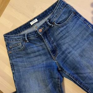 Bootcut Jeans från Lindex - Karen - Snygga blå jeans från Lindex (Karen) med klassisk femficksdesign och bootcut-ben. Jeansen har en mörkblå tvätt med lätt slitning och sitter tajt över höfterna innan de släpper ut vid vaden. Perfekta för dig som gillar en tidlös och trendig look.