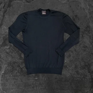 Svart stickad tröja Oscar Jacobson - En klassisk svart stickad tröja från Oscar Jacobson. Tröjan har rund halsringning, ribbade muddar och lång ärm. Perfekt för lager-på-lager och enkel att matcha med olika outfits.