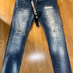 Dsquared2 blå jeans - Säljer ett par blå jeans som är märket Dsquared2 med coola slitningar och blekta detaljer. Modellen är denim med normal passform och klassisk femficksdesign. Jeansen har en svart Dsquared2-bälte och tydliga logotaggar. 