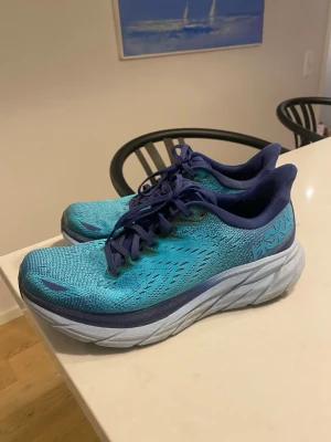 Blå Hoka Clifton 8 löparsko. - Säljer ett par Hoka sneakers i blå och turkos färg med tjock, vit sula och snörning. Ovandelen är i ventilerande syntetmaterial med mönstrade detaljer och Hoka-logga på sidan. Perfekta för dig som gillar att springa eller gå långt.