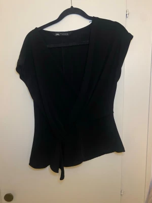 Svart omlottopp från Zara - Säljer en svart omlottopp från Zara med knyt framtill. Toppen har korta ärmar och en v-ringad design som ger en snygg siluett. Materialet är mjukt och lätt, perfekt för dig som gillar stilrena och enkla plagg. Passar dig som vill ha en klassisk look.