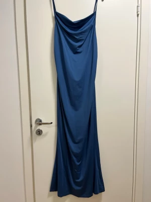 Blå långklänning med smala axelband - Säljer en elegant blå långklänning med smala axelband och draperad halsringning. Klänningen har en slank siluett och är tillverkad i ett mjukt, följsamt material som ger ett snyggt fall. Perfekt för dig som gillar stilrena och enkla plagg med modern touch.
