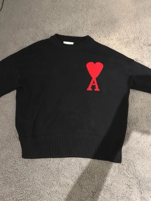 Svart stickad tröja från Ami - Svart stickad tröja från Ami med röd hjärtdetalj och bokstaven A på bröstet. Tröjan har ribbad hals, muddar och nederkant, samt en loose passform. Perfekt för dig som gillar stilrena plagg med en twist.