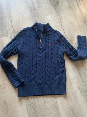 Marinblå stickad tröja Polo Ralph Lauren - Säljer en marinblå stickad tröja från Polo Ralph Lauren med kabelstickat mönster och röd broderad logga på bröstet. Tröjan har lång ärm, ribbad krage och half zip med läderdetalj. Perfekt för dig som gillar klassisk och stilren look.