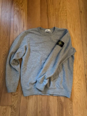Grå sweatshirt från Stone Island M - Säljer en grå sweatshirt från Stone Island i storlek M. Tröjan har rund hals, ribbade muddar och den klassiska Stone Island-loggan på vänster ärm. Tillverkad i mjuk bomull som är skön att ha på sig. Perfekt för dig som gillar stilrena och ikoniska plagg.