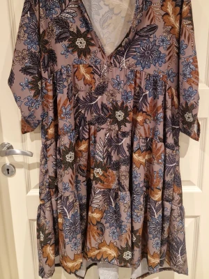 Blommig midiklänning från Zanca Sonne - Säljer en flowy midiklänning från Zanca Sonne med blommigt mönster i blått, brunt och svart på en ljuslila botten. Klänningen har v-ringning, trekvartsärm med knappdetalj och volang i kjolen. Perfekt för dig som gillar boho-vibe och färg!