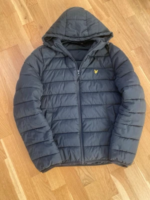 Svart pufferjacka från Lyle & Scott - Säljer en svart pufferjacka från Lyle & Scott med huva och gul logga på bröstet. Jackan har horisontella quiltade paneler och dragkedja framtill. Perfekt för kyliga dagar och enkel att matcha med olika outfits.