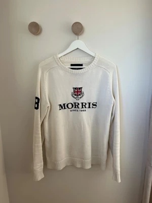 Vit stickad tröja från Morris - Snygg vit stickad tröja från Morris i storlek medium. Tröjan har broderad logga och text på bröstet samt siffran 68 i svart på ena ärmen. Rund hals och ribbade muddar vid ärmslut och nederkant. Perfekt för dig som gillar klassisk stil. OBS! Har ett par fläckar längre ner på tröjan, syns däremot inte särskilt mycket vid användning.