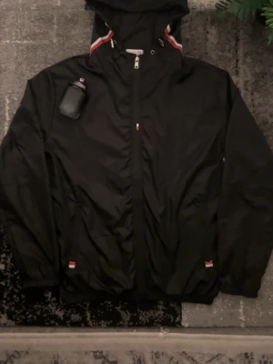 Svart vindjacka från Moncler - Svart vindjacka från Moncler med huva och dragkedja framtill. Jackan har röda och vita detaljer vid huvan och muddar, samt Moncler-logga på ärmen. Materialet är lätt och syntetiskt, perfekt för blåsiga dagar. Snygg och sportig look med normal passform.