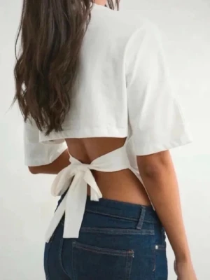 Vit croppad topp med knyt i ryggen - Säljer en vit croppad topp med öppen rygg och breda korta ärmar. Toppen har en snygg knytning baktill som ger en cool och trendig look. Perfekt för varma dagar och att styla med jeans eller kjol.