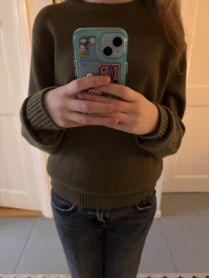 Olivgrön stickad tröja med rund hals - Säljer en olivgrön stickad tröja med rund hals och ribbade muddar vid ärmslut och nederkant. Tröjan har en enkel och klassisk design med lång ärm och normal passform, perfekt för lager-på-lager under kyliga dagar.