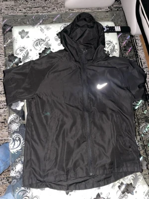 Svart Nike vindjacka med huva - Nike vindjacka i svart med huva och hel dragkedja framtill. Jackan har två sidofickor med dragkedja och reflekterande Nike-logga på bröstet. Lätt och smidig design, perfekt för löpning och utomhusaktiviteter. Materialet är polyester och insidan har Nike Running-detaljer.
