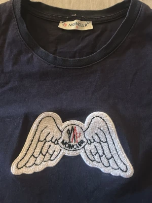 Mörkblå Moncler t-shirt  - Mörkblå t-shirt från Moncler med rund hals och ett stort broderat vingmärke på bröstet. T-shirten är i mjuk bomull och har en klassisk passform med korta ärmar. Perfekt för dig som gillar stilrena plagg med en unik detalj.      Hör av er vid frågor och funderingar.