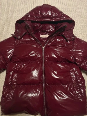 Vinröd pufferjacka från Nelly - Säljer en glansig vinröd pufferjacka från Nelly med huva och dragkedja framtill. Jackan är helt ny och har en oversized look, perfekt för kalla dagar. Fickor på sidorna och hög krage för extra värme. ☺️☺️