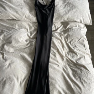 Svart medellång satinklänning med smala band - Supersnygg svart långklänning i satin med tunna axelband som korsas i ryggen och en draperad urringning. Klänningen har en slimmad passform och ett elegant, minimalistiskt uttryck. Perfekt för dig som vill ha en stilren och trendig look.