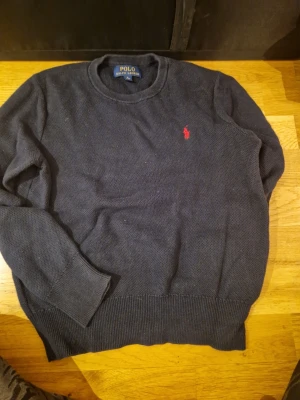 Mörkblå stickad tröja Polo Ralph Lauren - Snygg mörkblå stickad tröja från Polo Ralph Lauren i storlek M 10-12 år. Klassisk rund hals och långärmad modell med röd broderad logga på bröstet. Ribbstickade muddar vid ärmslut och nederkant ger en schysst passform. Perfekt för dig som gillar stilrena plagg.