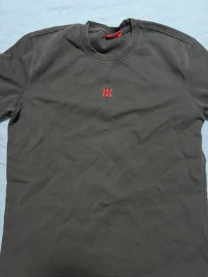 Svart t-shirt från Hugo med röd logga - Svart t-shirt från Hugo med rund hals och korta ärmar. På bröstet finns en liten röd broderad Hugo-logga som ger en clean och stilren vibe. Materialet är mjuk bomull som känns skönt mot huden. Perfekt för dig som gillar minimalistisk design och snygga detaljer.