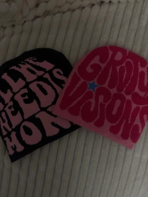 Två färgglada mössor med text - Säljer två stickade mössor med coola, stora texter i retrostil. Ena mössan är svart med rosa text och den andra är rosa med röd text och en liten blå stjärna. Perfekt för dig som vill sticka ut med färg och grafisk design. Båda för 180kr 