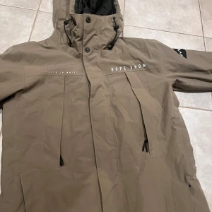 Beige skidjacka från Dope Snow - Säljer en beige skidjacka från Dope Snow med huva och snygga svarta detaljer. Jackan har dragkedjor, snörning vid huvan och tryckt logga på bröstet samt patch på ärmen. Perfekt för dig som gillar street och outdoor vibes.