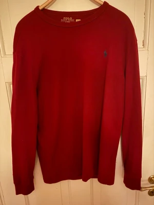 Röd långärmad tröja från Polo Ralph Lauren - Säljer en stilren röd långärmad tröja från Polo Ralph Lauren med klassisk rund hals och diskret blå logga broderad på bröstet. Tröjan är i mjuk bomull och har en normal passform, perfekt för dig som gillar enkel och snygg design.