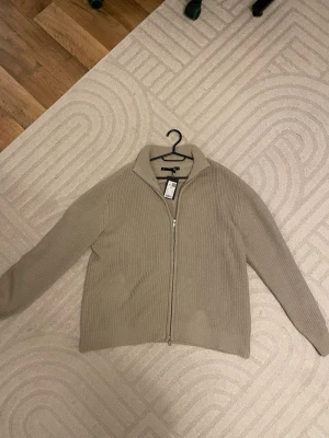 Beige stickad kofta med dragkedja  - Säljer en beige stickad kofta från 157% i storlek M. Koftan har ribbad struktur, hög krage och hel dragkedja framtill. Perfekt för lager-på-lager och chill dagar. Enkel och stilren design som funkar till det mesta.