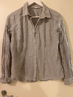 Randig skjorta från HM , storlek 44 - Snygg skjorta från HM  i storlek 44 med klassiskt svartvitt randigt mönster. Skjortan har lång ärm, knappar framtill och en krage. Tillverkad i mjuk bomull som känns skön mot huden. Perfekt för dig som gillar stilrena och tidlösa plagg.