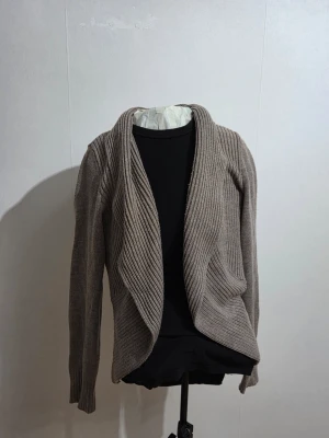 Beige stickad kofta från H&M - Säljer en beige stickad kofta från H&M Basic i storlek M. Koftan har ett öppet och omlottliknande fall framtill, ribbad struktur och långa ärmar. Perfekt att slänga över en t-shirt eller linne när det är lite kyligt ute.