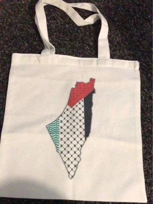Palestina Vit shoppingväska med tryck - Säljer en vit shoppingväska i bomull med ett stort tryck i mitten som föreställer Palestina  karta med olika mönster och färger i rött, svart, grönt och vitt. Tygväska Vita Kvinnor Män Handväska  Produktstorlek: 380,00 x 340,00 x 20,00 MM/14,96 x 13,39 x 0,79 tum  Form: Vanlig tygväska med stor kapacitet  Huvudmaterial: Polyester Bomull .Väskan har långa handtag och är perfekt för att bära böcker, mat eller andra grejer.  