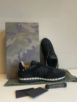 Valentino Rockrunner svarta sneakers - Svarta Valentino Rockrunner sneakers med coola nitar runt hälen och sulan. Skorna har en blandning av mocka och textil, snörning och en grov platt sula. Kommer med originalkartong i camouflage och dustbag. Perfekt för dig som gillar edgy designerstil.