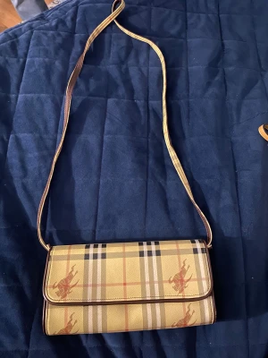 Rutig axelväska från Burberry - Säljer en klassisk axelväska från Burberry med det ikoniska rutiga mönstret i beige, brunt och svart. Väskan har en lång axelrem med bruna detaljer och insida med Burberry-logga. Materialet är skinn och textil, perfekt för dig som gillar tidlös design.