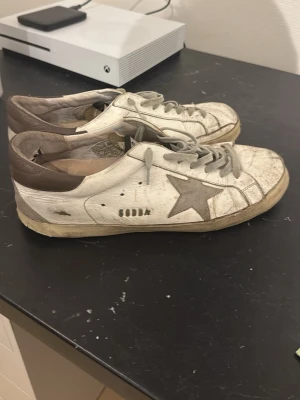 Golden Goose sneakers  - Tja säljer ett par Golden Goose sneakers i vitt skinn med grå stjärndetalj på sidan och grå snörning. Skorna har beige och bruna toner på hälpartiet och platt sula. Klassisk låg modell med rund tå och slitna detaljer för en cool look. Skorna är använda mycket, går dock att fräscha upp dom om man vill!Box och påse till skorna medföljer. Hör av er vid eventuella frågor!