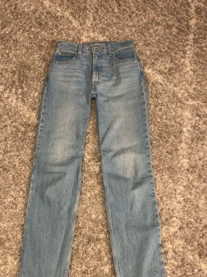 Blå mid waist slim straight jeans - Säljer ett par ljusblå Levi's jeans i modellen 70's High Slim Straight. De har medel midja, raka ben och klassisk femficksdesign. Jeansen är gjorda i jeansmaterial med snygga slitningar och detaljerade sömmar. Perfekta för dig som gillar retrostil och bekväma passformer. Lite tighta vid låren