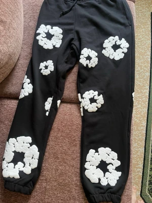 Svarta mjukisbyxor Denim Tears med blommor - Svarta mjukisbyxor från Denim Tears med stora vita blommiga mönster över hela byxorna. Byxorna har resår vid bensluten och snörning i midjan. Materialet är mjukt och bekvämt, perfekt för chill dagar. Snygg och unik design som sticker ut.