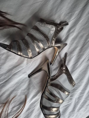 Silvriga klackar med ormskinnsdetaljer - Säljer ett par unika pumps från Sthef med silvriga ormskinnsmönstrade remmar och transparent mesh. Skorna har öppen tå, hög klack och justerbart spänne vid ankeln. Perfekt för dig som vill sticka ut med coola detaljer och metallic vibes. Kan pruta på priset. NÄSTAN NYA