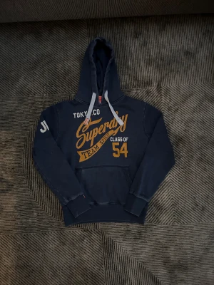 Mörkblå Superdry hoodie - Mörkblå hoodie från Superdry med stort gult och vitt tryck på bröstet och ärmen. Klassisk känguruficka, huva med vita snören och ribbade muddar. Perfekt för dig som gillar streetstyle och sportig vibe.