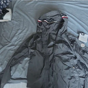 vindjacka från Moncler med huva - vindjacka från Moncler med huva och snygga snörningsdetaljer. Jackan har dragkedja framtill, Moncler-logga på ärmen och en diskret rödvit rand på insidan av huvan. Perfekt för dig som gillar stilren design och exklusiva märken. Den är helt ny