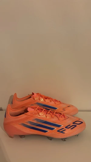 Adidas F50 CORAL BLAZE Elite  - Säljer ett par Adidas F50 fotbollsskor i en riktigt orange färg med blå detaljer och vita linjer. Skorna har snörning, platt sula med dobbar och en smal, aerodynamisk form. Tillverkade i syntetmaterial för lätt känsla och maxad speed på planen.