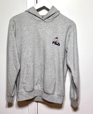 Grå hoodie från FILA med blomtryck - Grå hoodie från FILA med snyggt tryck av röda blommor och logga på bröstet. Tröjan har huva, magficka och långa ärmar med ribbade muddar. Perfekt för chill dagar och enkel att matcha med jeans eller joggers.