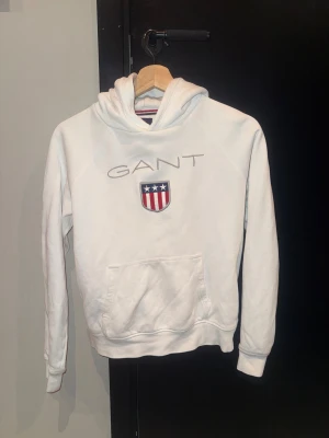 Vit hoodie från GANT med logga - Vit hoodie från GANT med broderad logga och sköld på bröstet. Tröjan har huva, magficka och långa ärmar. Mjuk och skön i bomullsmaterial, perfekt för chill dagar. Passar dig som är 13-14 år och gillar klassisk stil.