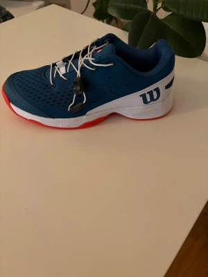 Wilson Rush Pro JR 4.0 QL blå/röd - Snygga Wilson Rush Pro JR 4.0 QL sneakers i blått med vita detaljer och röd sula. Skorna har ventilerande mesh, snörning och robust sula för bra grepp. Perfekta för dig som vill ha stilrena och sportiga tennisskor.