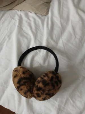 Leopardmönstrade öronmuffar - Snygga öronmuffar med leopardmönster i brunt och svart. De har en mjuk och fluffig pälsimitation som håller öronen varma och ett svart band som sitter bekvämt runt huvudet. Perfekt för att ge din vinterstil lite extra edge.