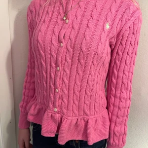 Rosa kofta från Polo Ralph Lauren - Supersöt rosa kofta från Polo Ralph Lauren med kabelstickat mönster, volangkant nertill och pärlemorknappar framtill. Klassisk logga broderad på bröstet. Långärmad och i mjuk bomull, perfekt för lager på lager under höst och vår. Strl 12-14 år som xxs dam