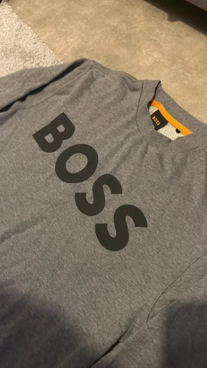 Grå sweatshirt från BOSS - Snygg grå sweatshirt från BOSS med stort svart logotryck över bröstet. Tröjan har rund hals, långa ärmar och ribbade muddar vid ärmslut och nederkant. Perfekt för dig som gillar stilrena och klassiska plagg med tydlig branding.