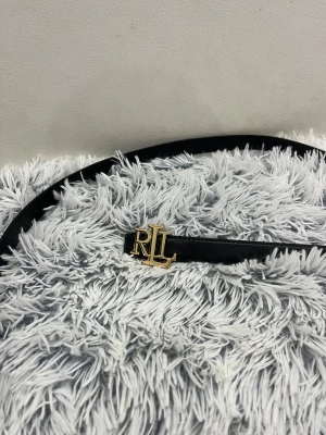 Svart skärp  - Snyggt svart bälte med ett stilrent RL-spänne i guld. Bältet är smalt och gjort i skinn, perfekt för att lyfta vilken outfit som helst med en touch av lyxig känsla.