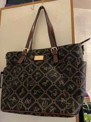 Svart handväska från Giulia Pieralli - Stilren svart handväska från Giulia Pieralli med bruna handtag och guldfärgade detaljer. Väskan har ett unikt mönster med kedjor och accessoarer i guld, samt logotypplatta framtill. Tillverkad i syntetmaterial och har flera fack och dragkedja.